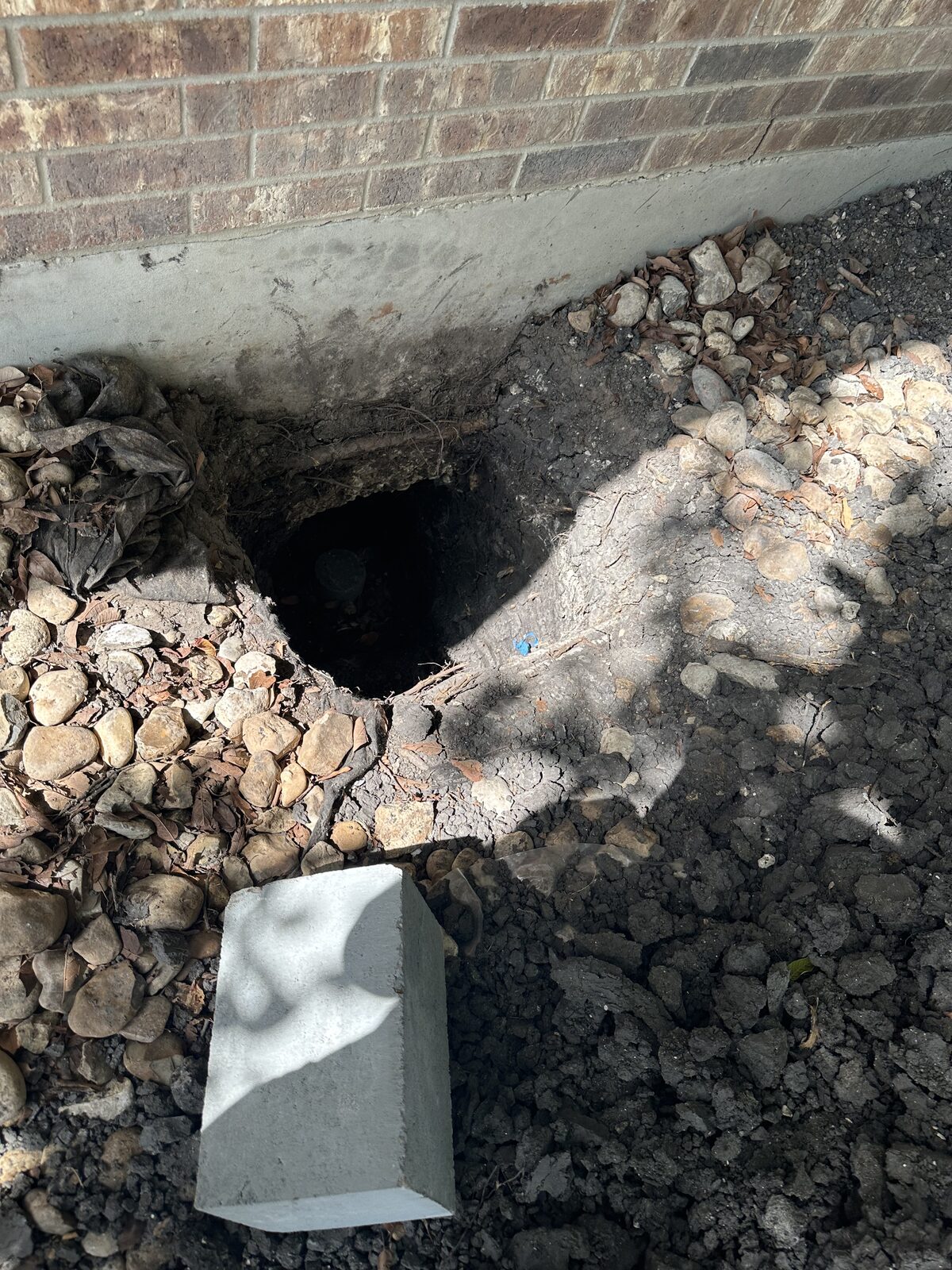 Partially backfilled pier hole in a river-rock bed along a brick home's foundation after pier installation