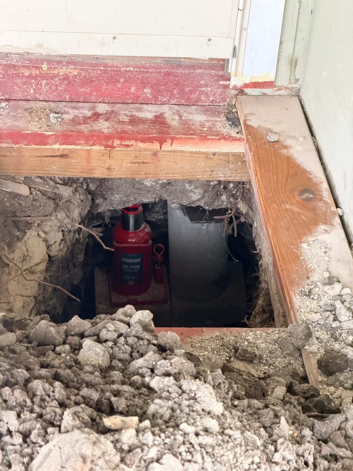 A red 20-ton bottle jack beside a finished hybrid pier stack inside a board-framed pier hole under a doorway