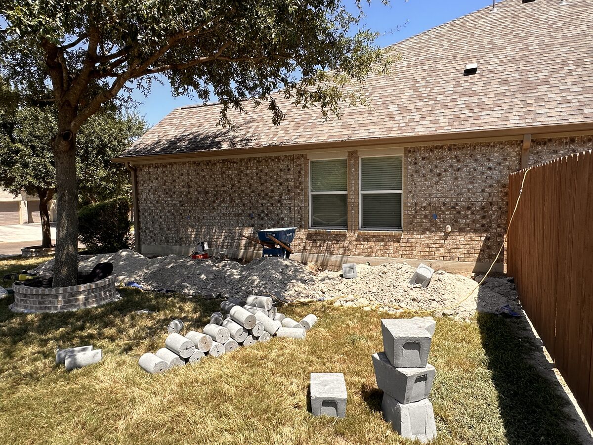 Brick home with pier holes and spoil along the rear foundation and concrete pilings staged on the lawn