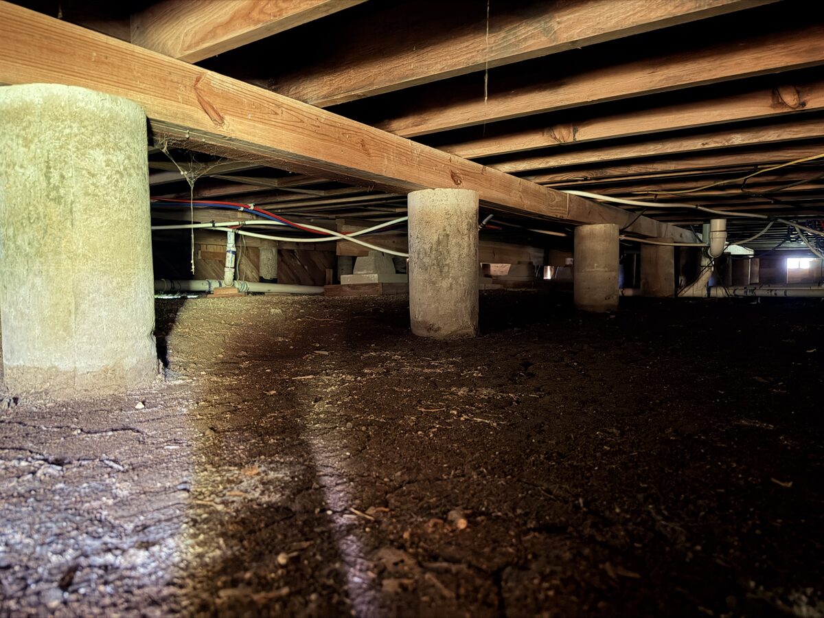 Concrete piers and wood beams supporting floor joists above a dirt crawl space floor