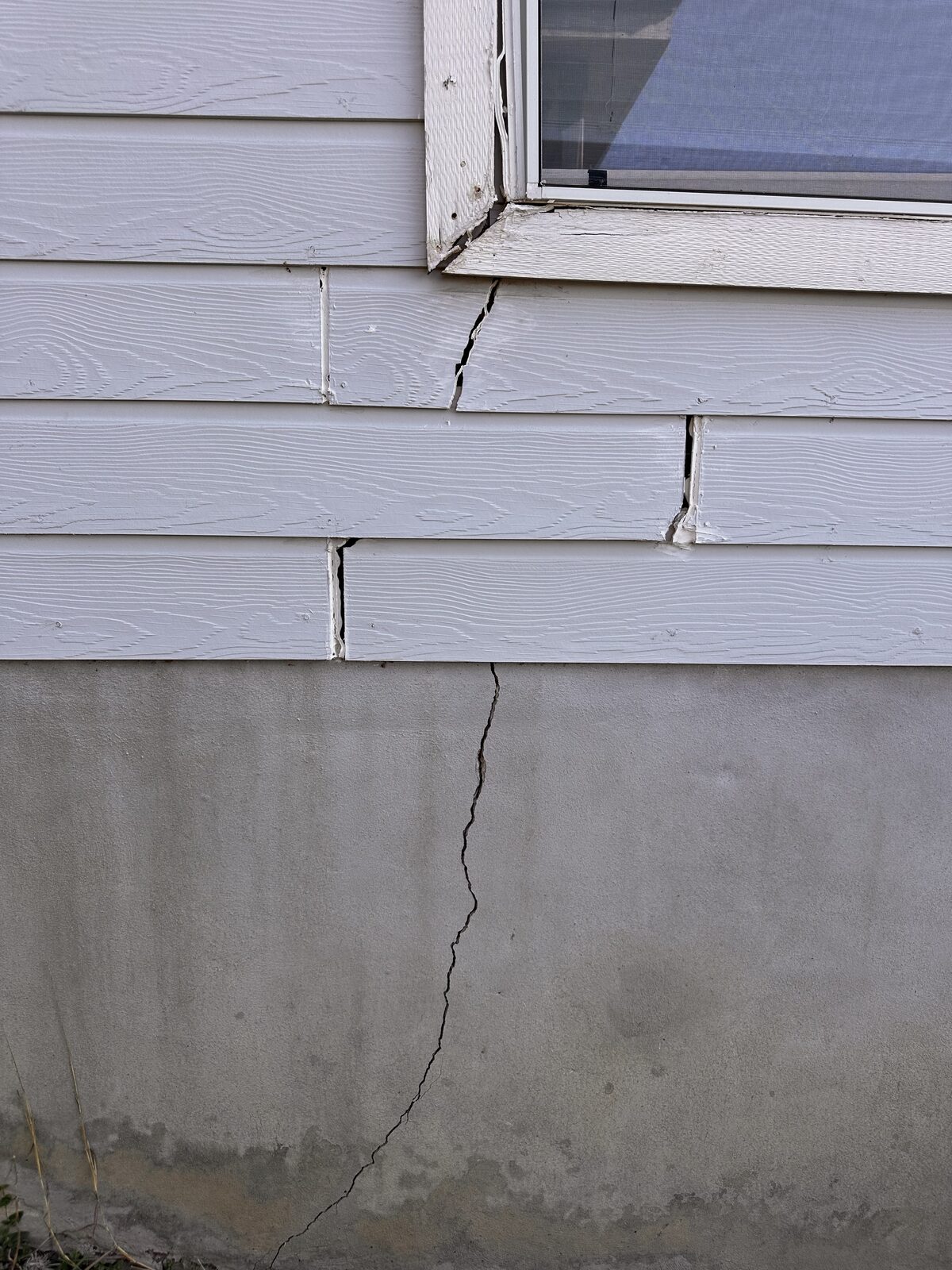 Foundation crack continuing up into separated siding boards below a window, showing structural movement
