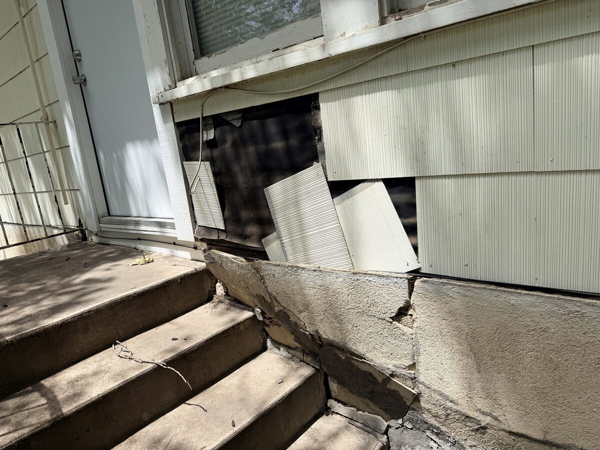 Porch steps beside a cracked stem wall and loose siding panels under a window