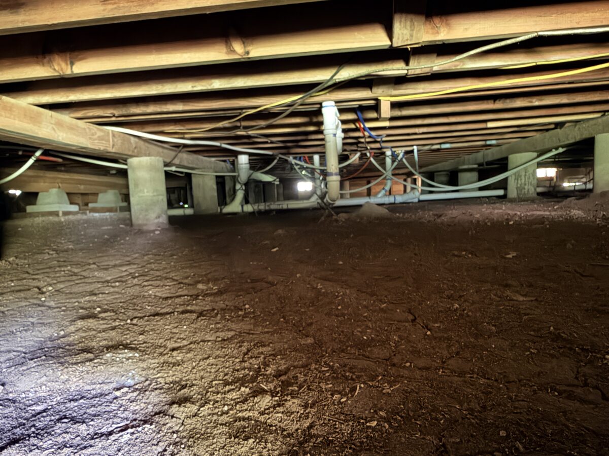 Wide crawl space view with concrete piers, plumbing lines, and floor joists overhead