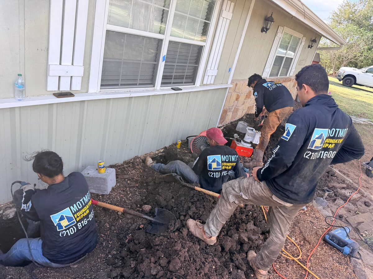 Crew in Motmot shirts digging hybrid pier holes along the front wall of a house with a pump and hammer nearby