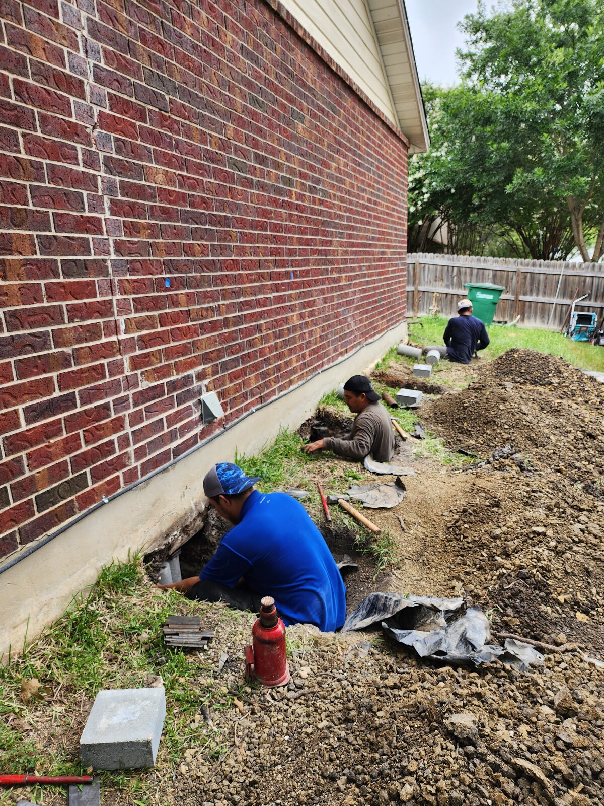Crew excavating a row of pier holes along a brick home's foundation during underpinning pier installation