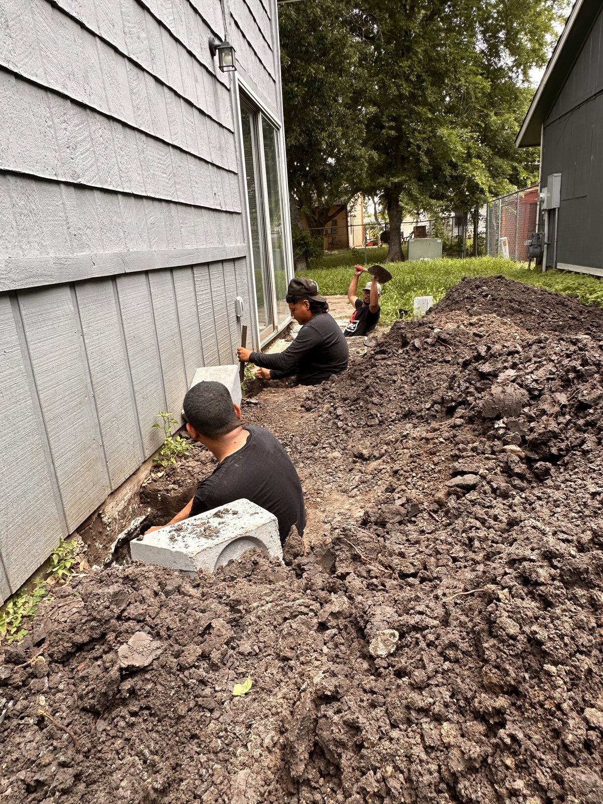 Three foundation repair crew members hand-digging pier pits along a gray-sided Central Texas home