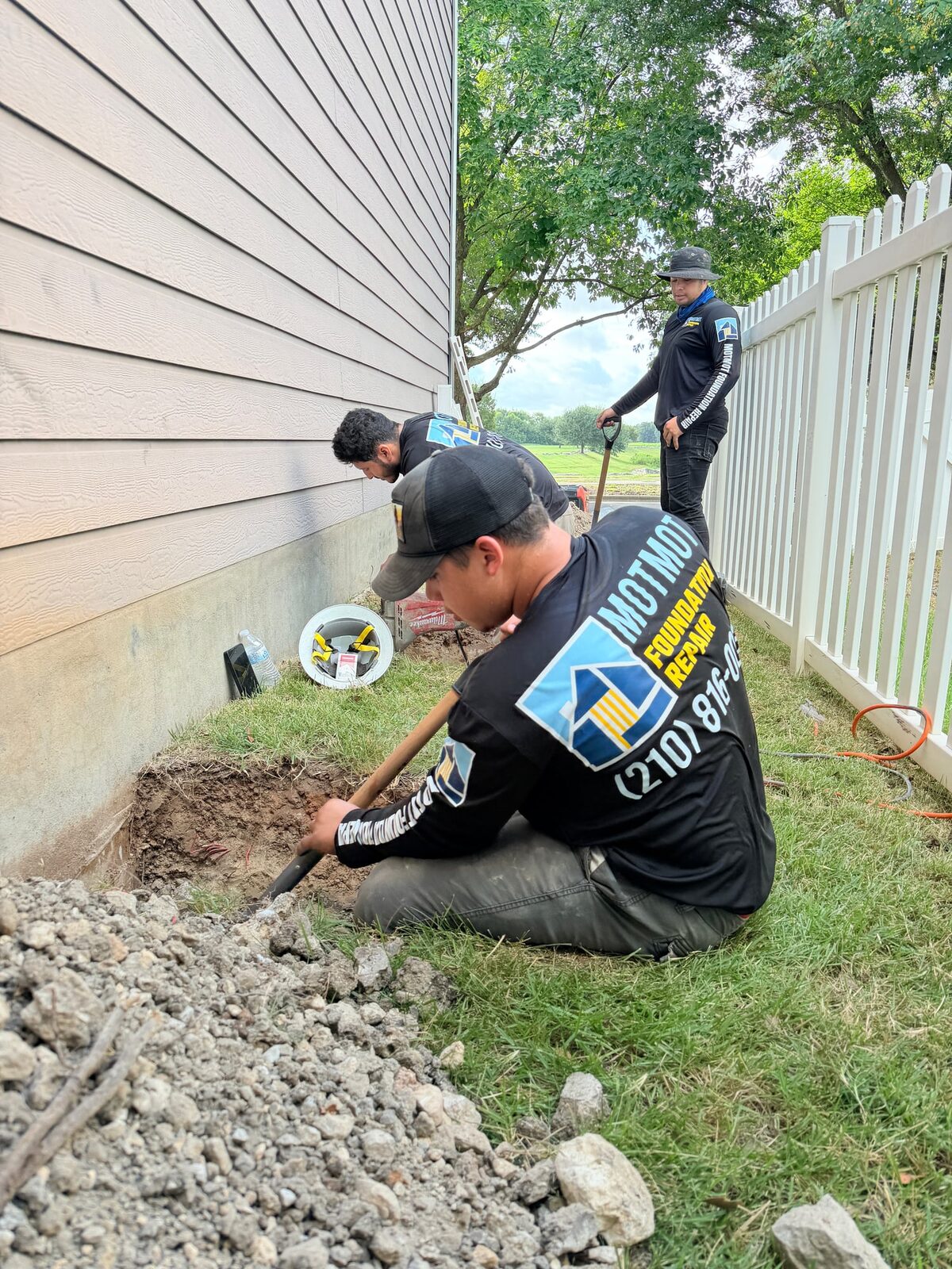 Three crew members digging a row of pier holes along a house, broken rock piled in front