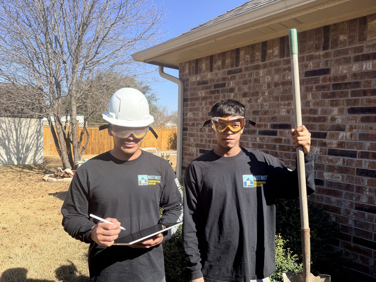 Two Motmot crew members with a tablet and shovel in front of a brick home