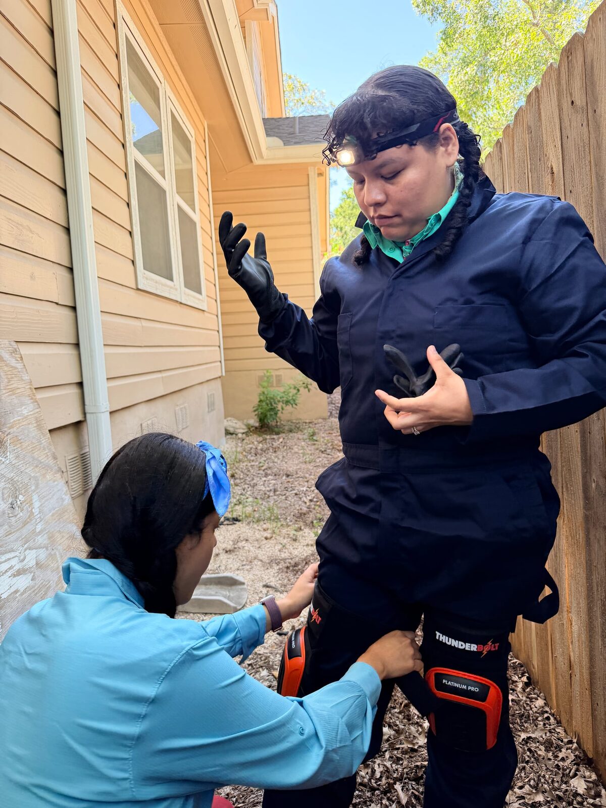 Crew member putting on coveralls, a headlamp and knee pads in a side yard, with louvered foundation vents in the wall behind