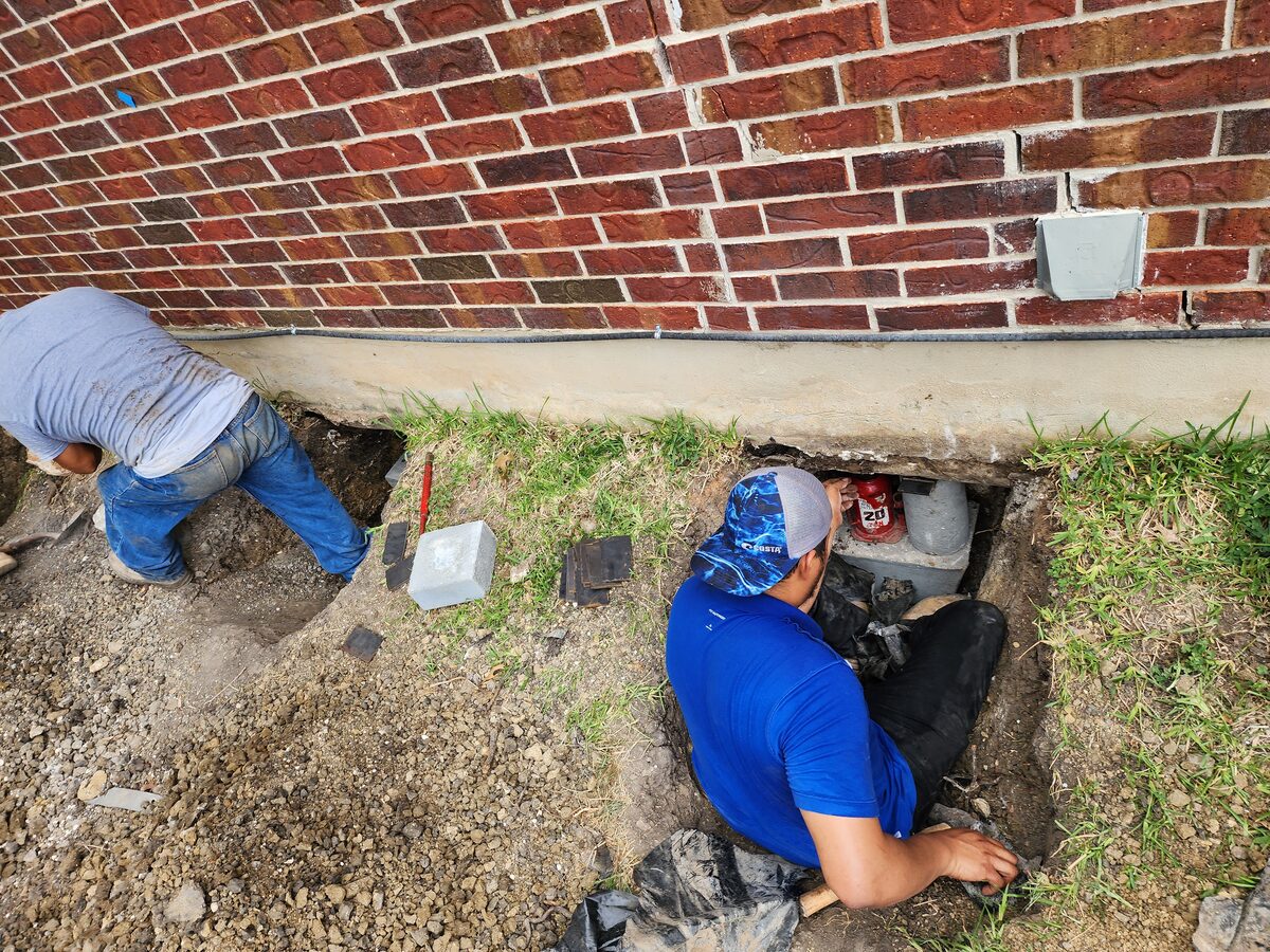 Foundation repair crew hand-digging pier pits along the brick exterior of a San Antonio area home