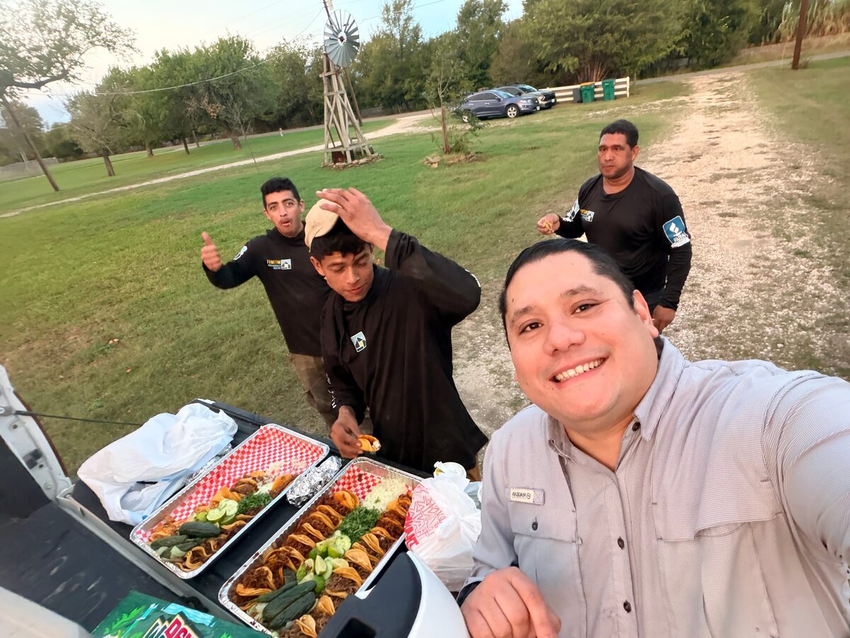 Smiling crew lead taking a selfie beside a pickup tailgate set with food trays, three crew members behind him, after a job in Belton, Texas