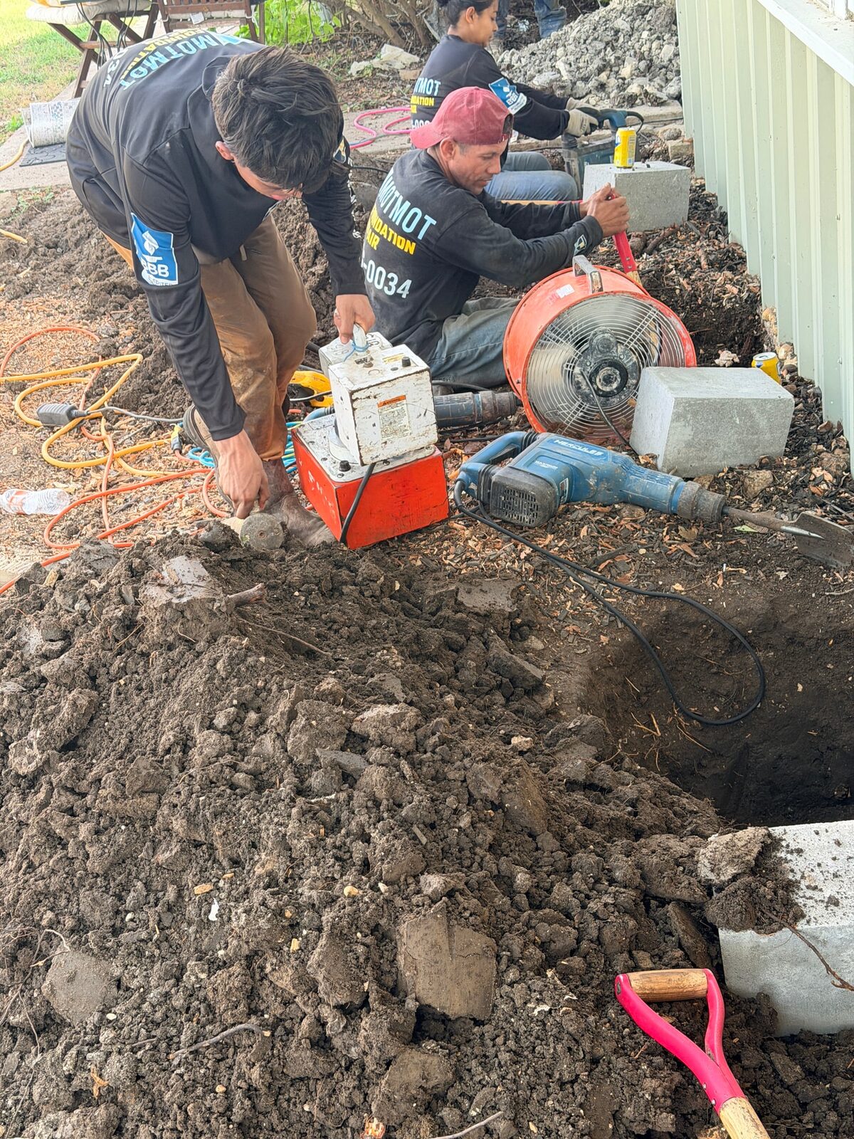 Two Motmot crew members setting up an electric hydraulic pump and a fan beside open pier holes, cap blocks stacked nearby