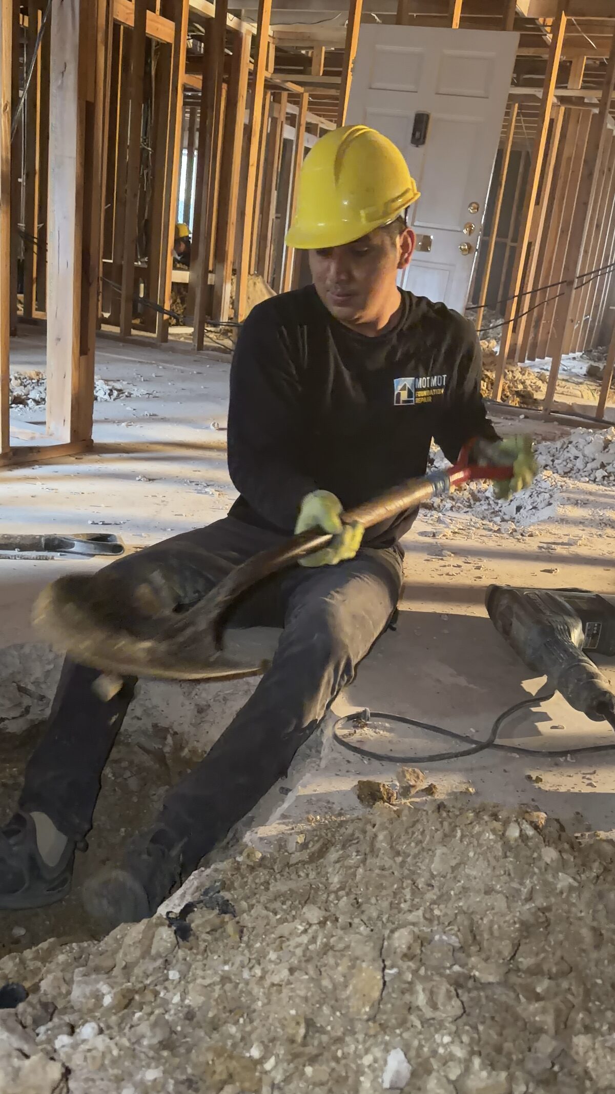 Foundation crew member in a hard hat shoveling soil from an interior pier hole inside a home under repair