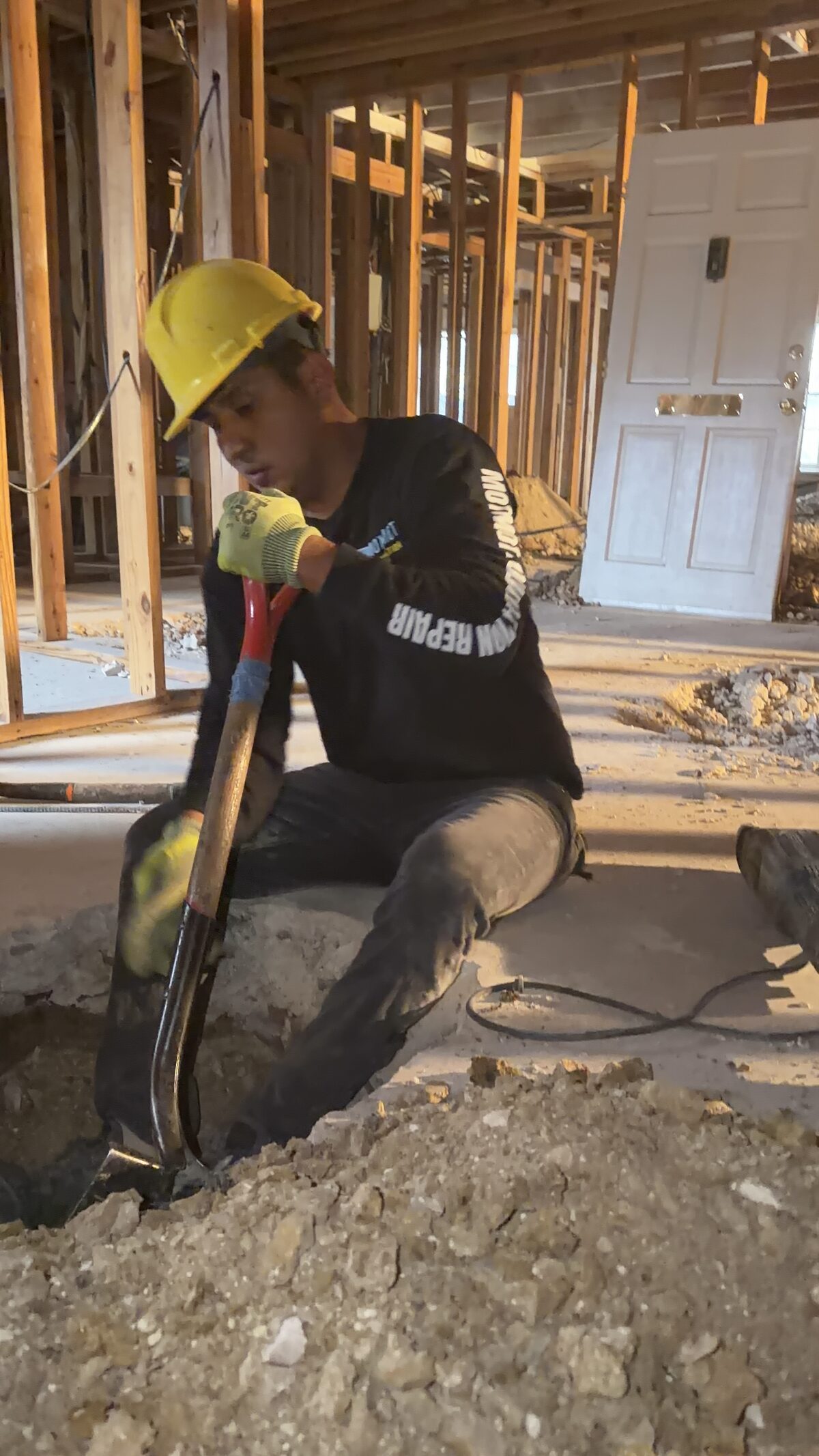 Crew member shoveling soil from a hand-dug interior pier hole during foundation repair inside a home