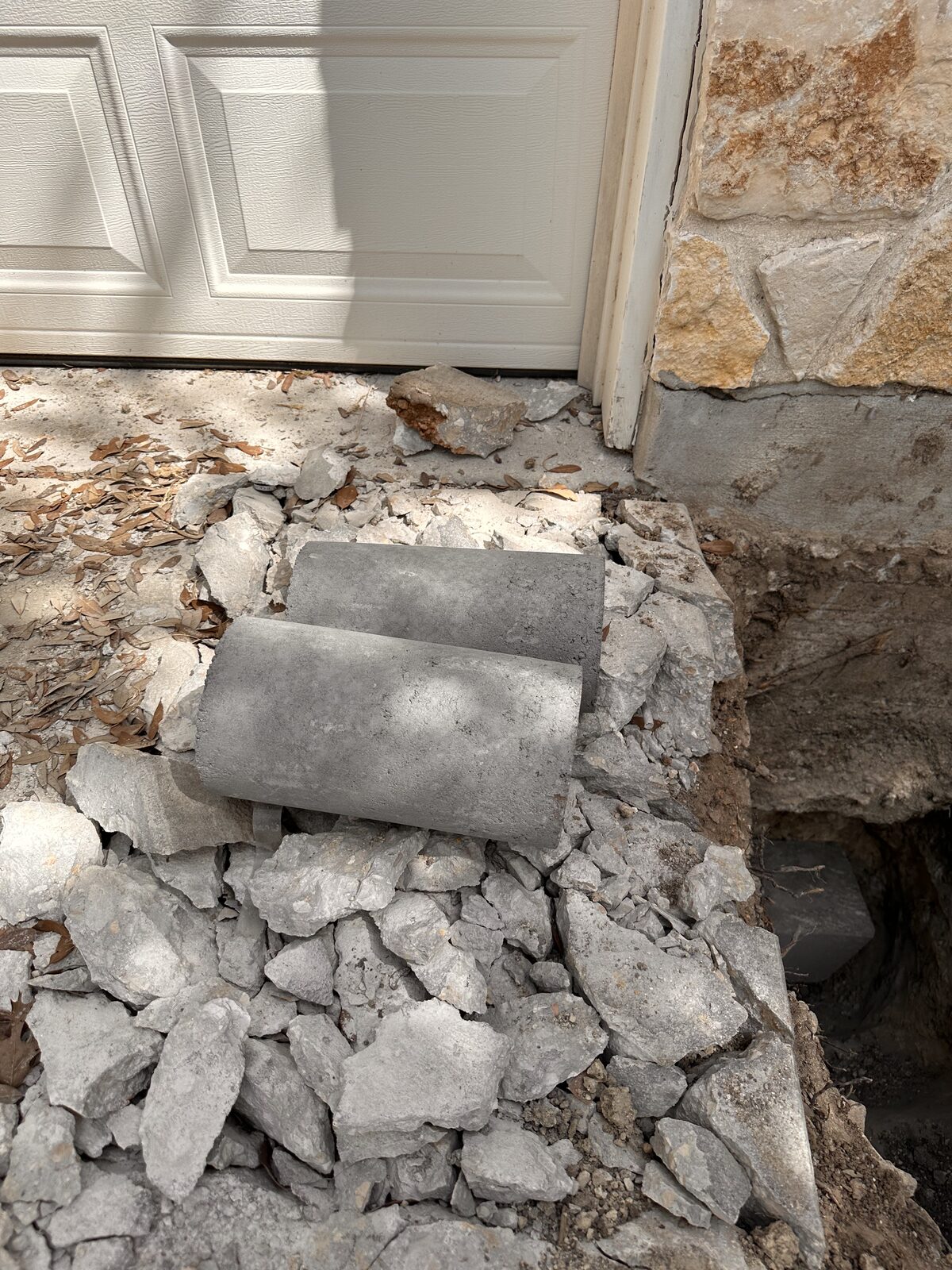 Cut concrete piling sections stacked on rock spoil beside a stone-veneer garage during foundation repair