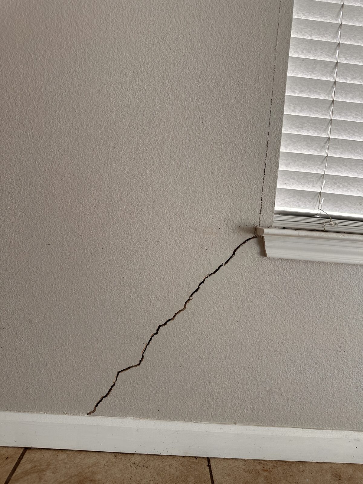 Diagonal settlement crack angling down the wall from a window corner with blinds