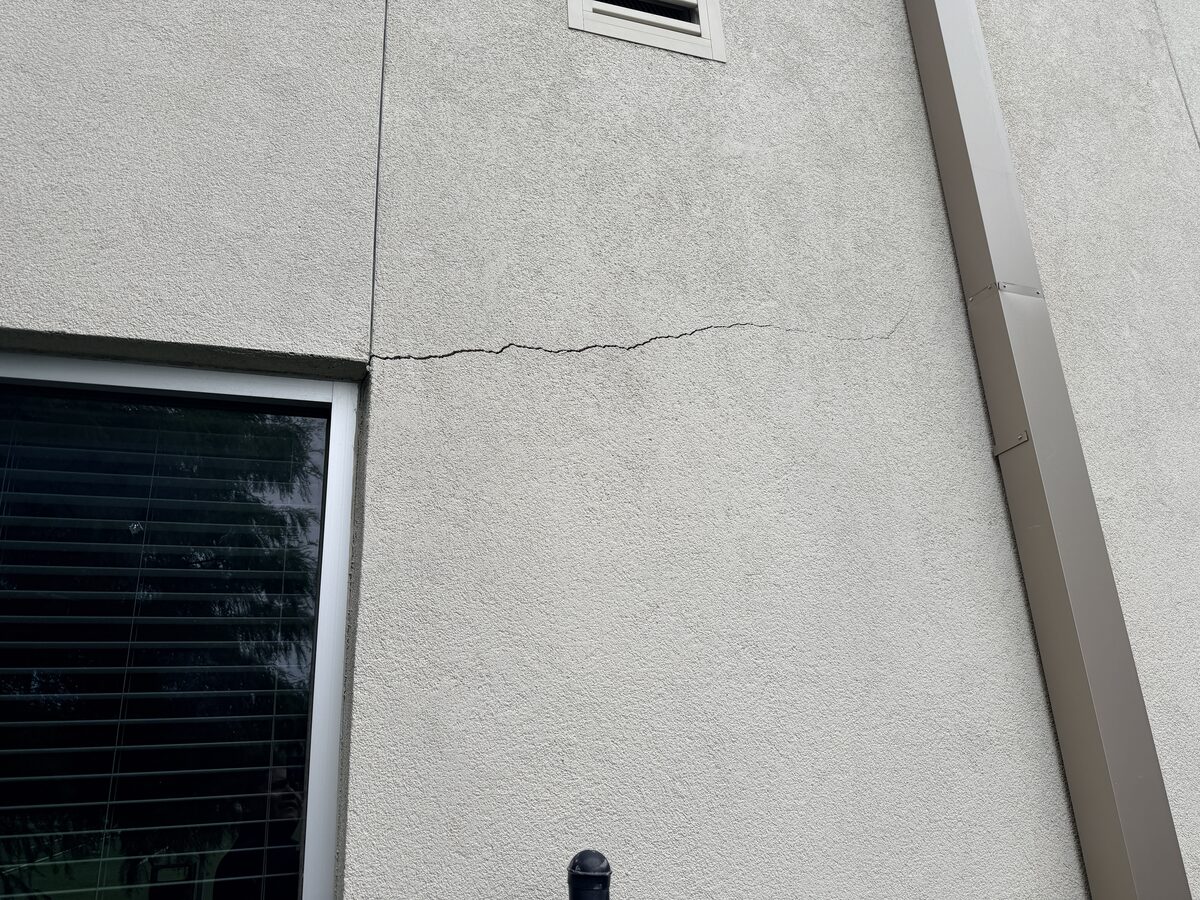 Crack running out of a window head across a light stucco wall next to a downspout on a commercial building