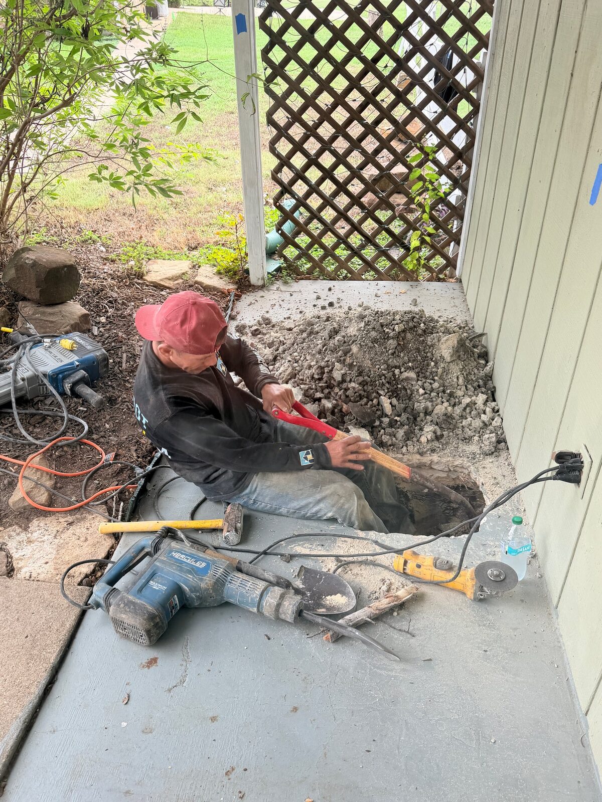 Crew member sitting in a hand-dug pier hole at the corner of a back porch slab, tools laid out on the porch