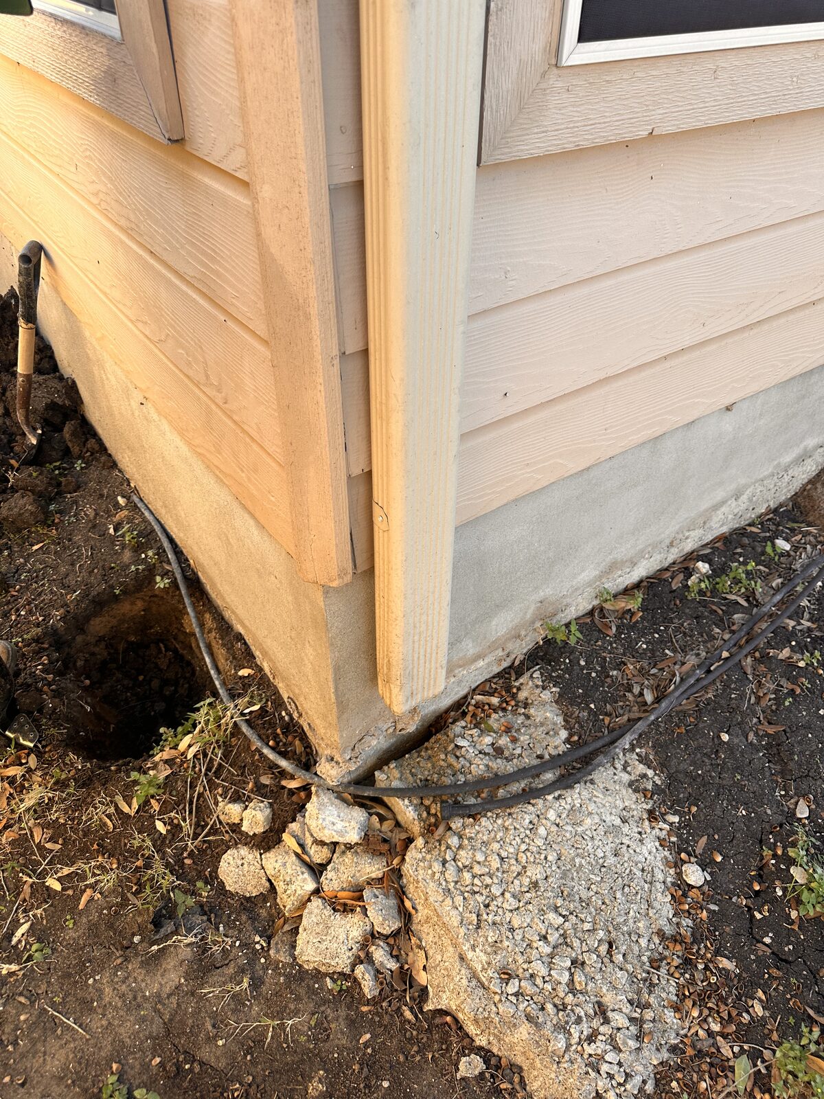 Excavated corner of a siding-clad home showing the exposed footing and gravel during pier work