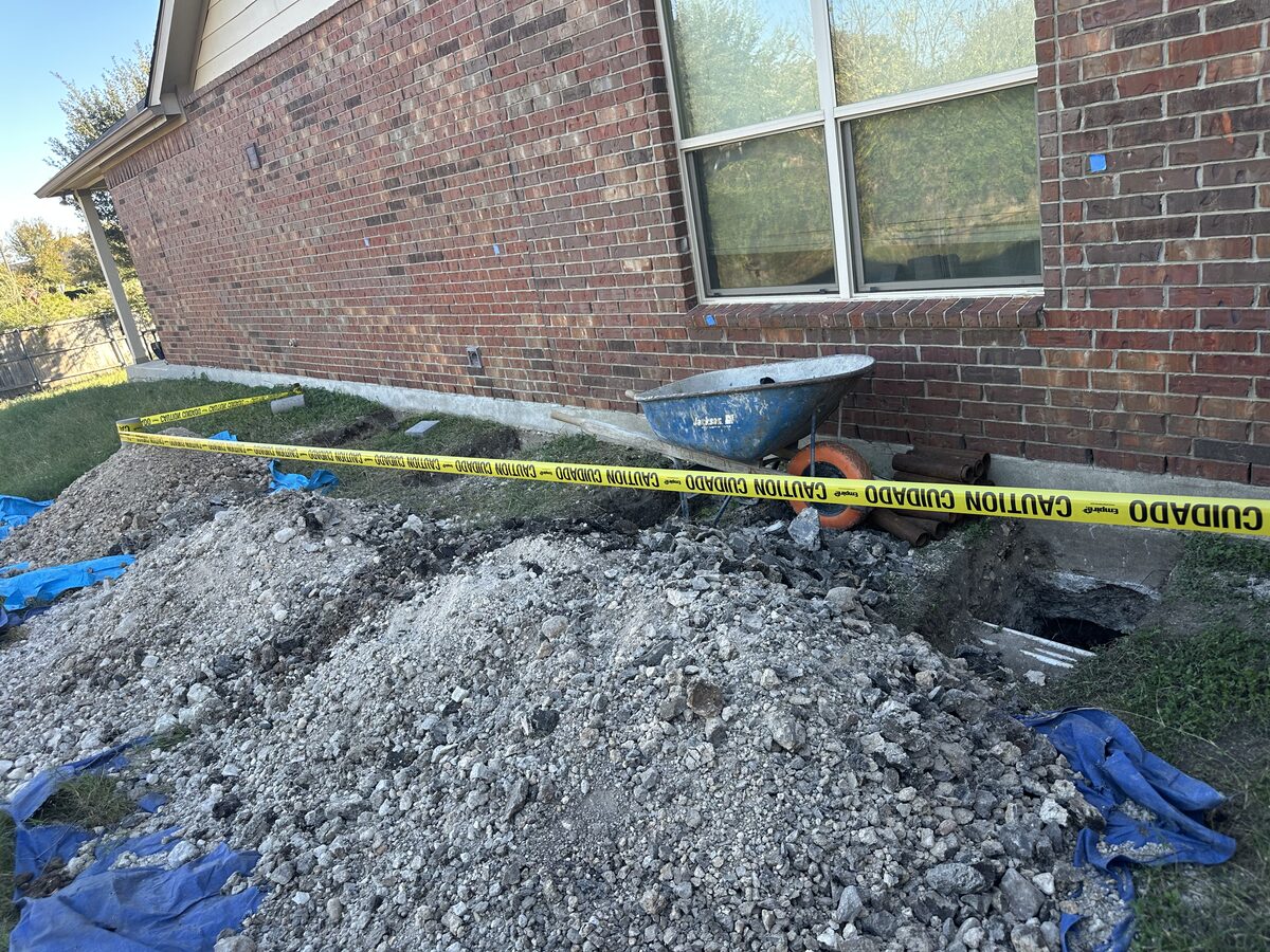 Excavated spoil pile with caution tape and a wheelbarrow beside pier holes along a brick home's foundation