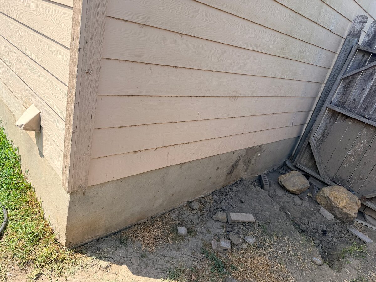Exposed concrete slab corner of a sided house with dry, cracked clay and loose rocks beside a fence
