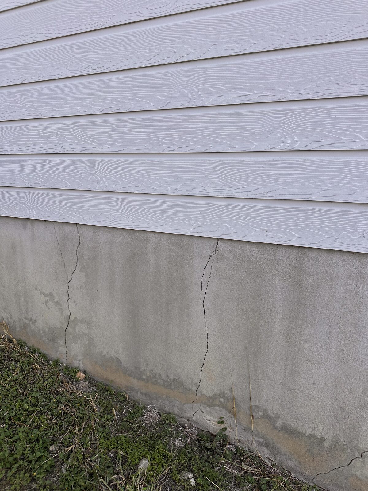Hairline crack in a concrete foundation wall at grade beneath light gray siding