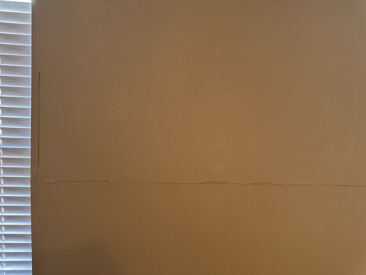 Long horizontal drywall crack across a bedroom wall beside a window, evidence of foundation movement