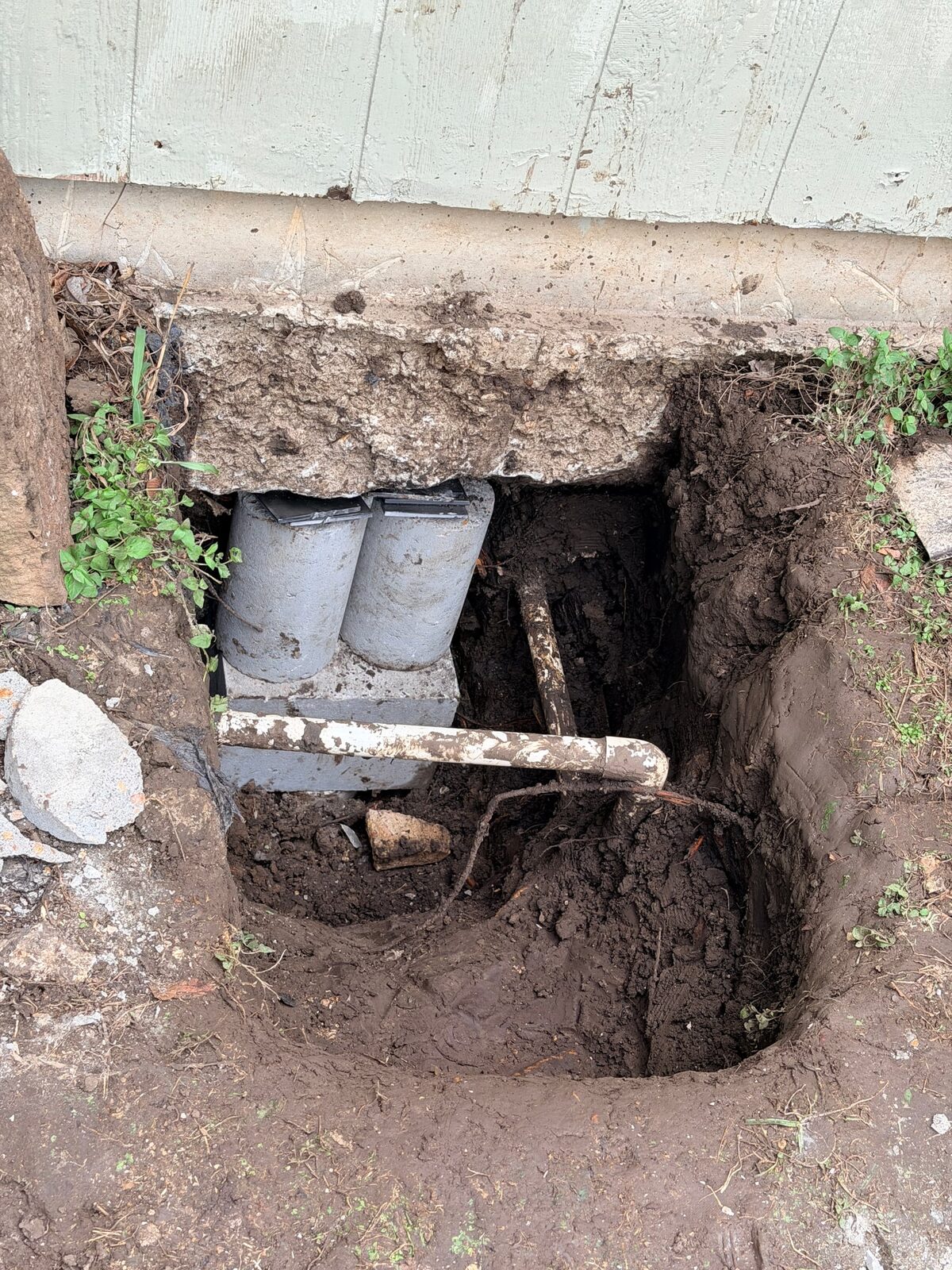 Hybrid pier in a hand-dug hole under a siding wall, two concrete cylinders on a block with steel shims under the beam and an old pipe crossing the hole