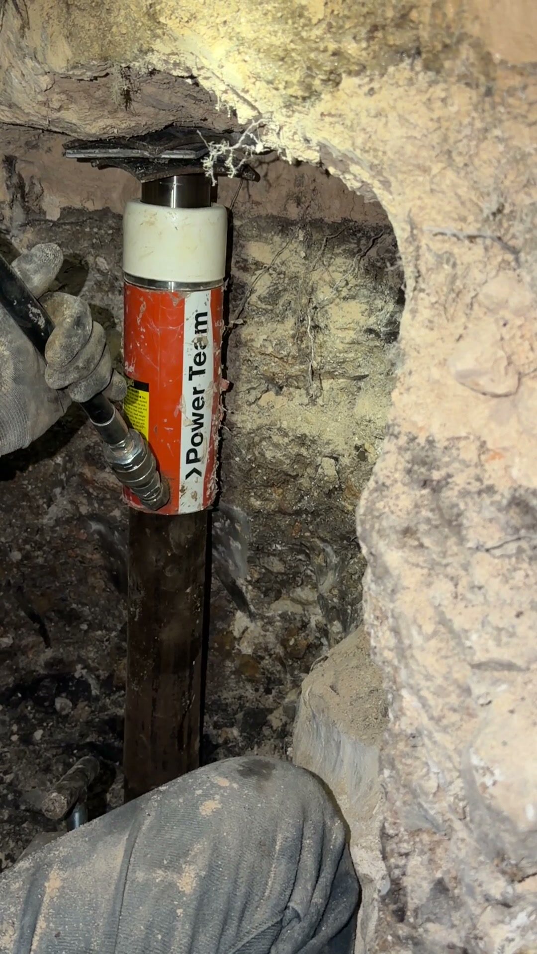 Hydraulic ram pressing a steel pier pipe straight down under the slab in a hand-dug tunnel