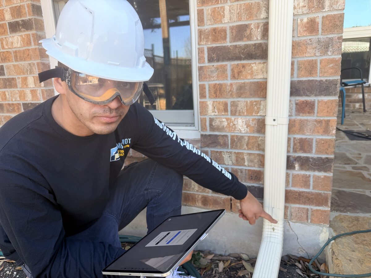 Inspector pointing to a hairline crack near brick weep holes while recording findings on a tablet