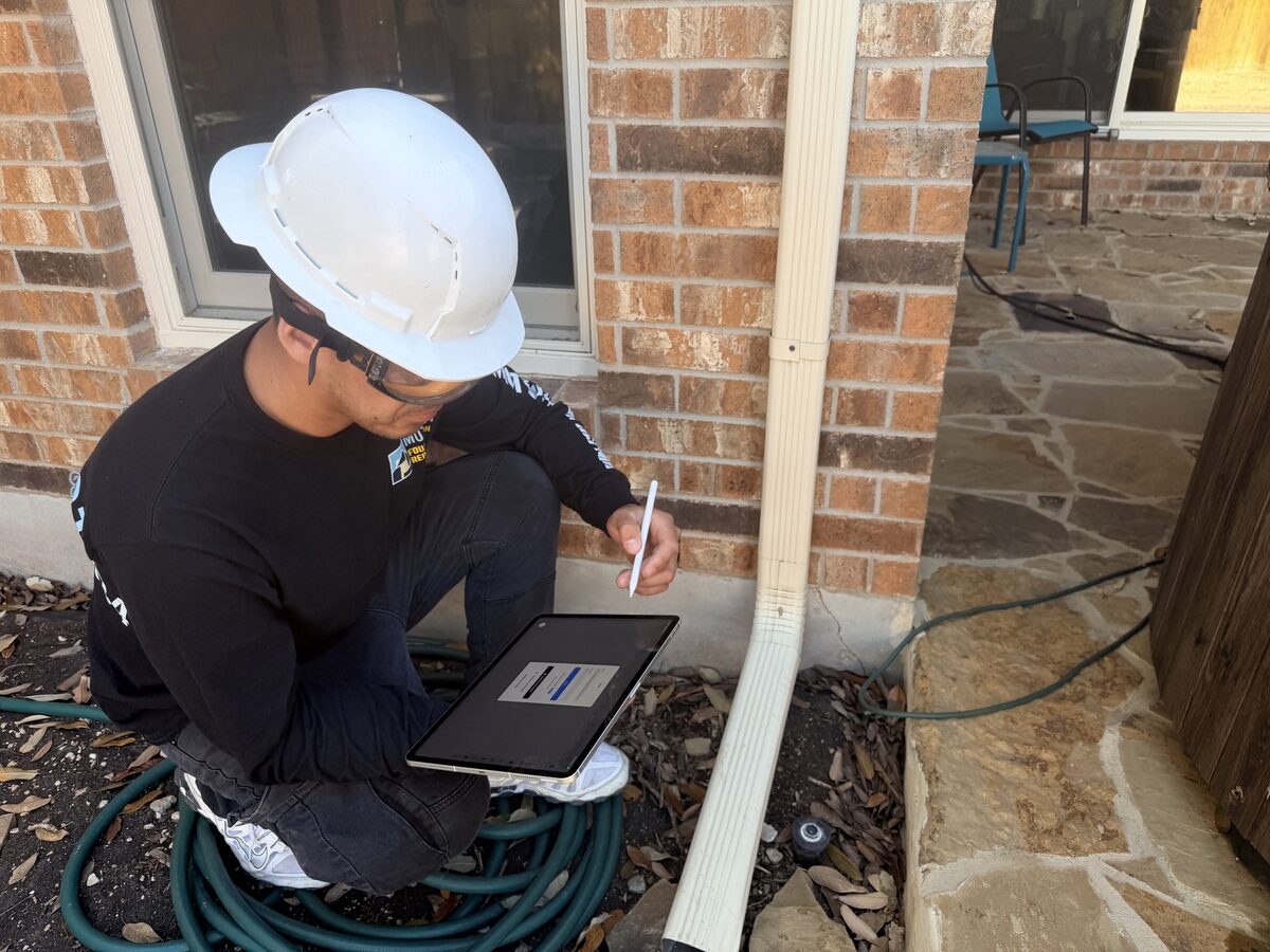Foundation inspector recording notes on a tablet beside a downspout at the slab edge of a brick home