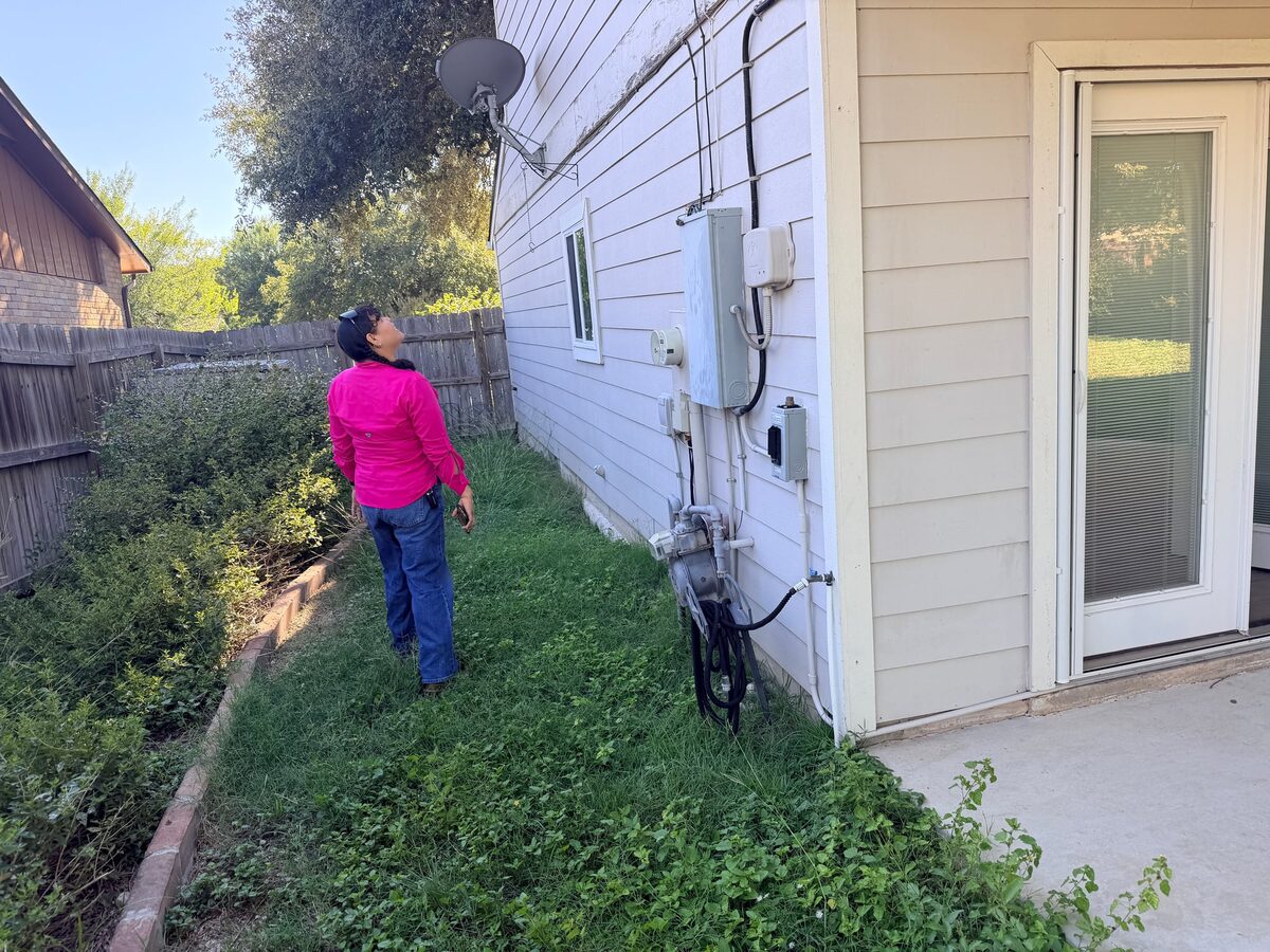 Inspector walking a grassy side yard and looking up along the siding, past a hose bib and the utility meters