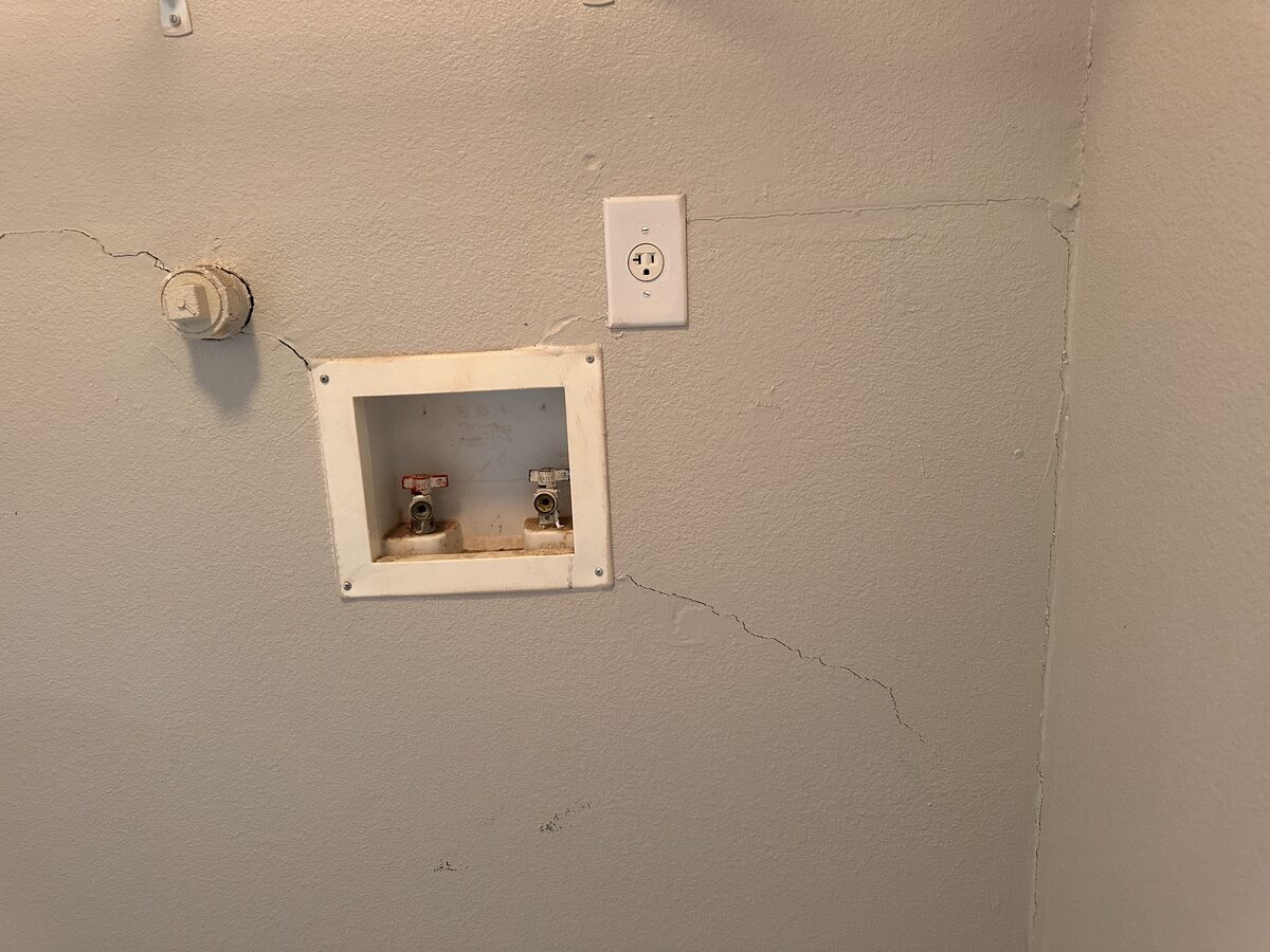 Diagonal drywall crack beneath a washing machine supply box in the laundry room of a settling home