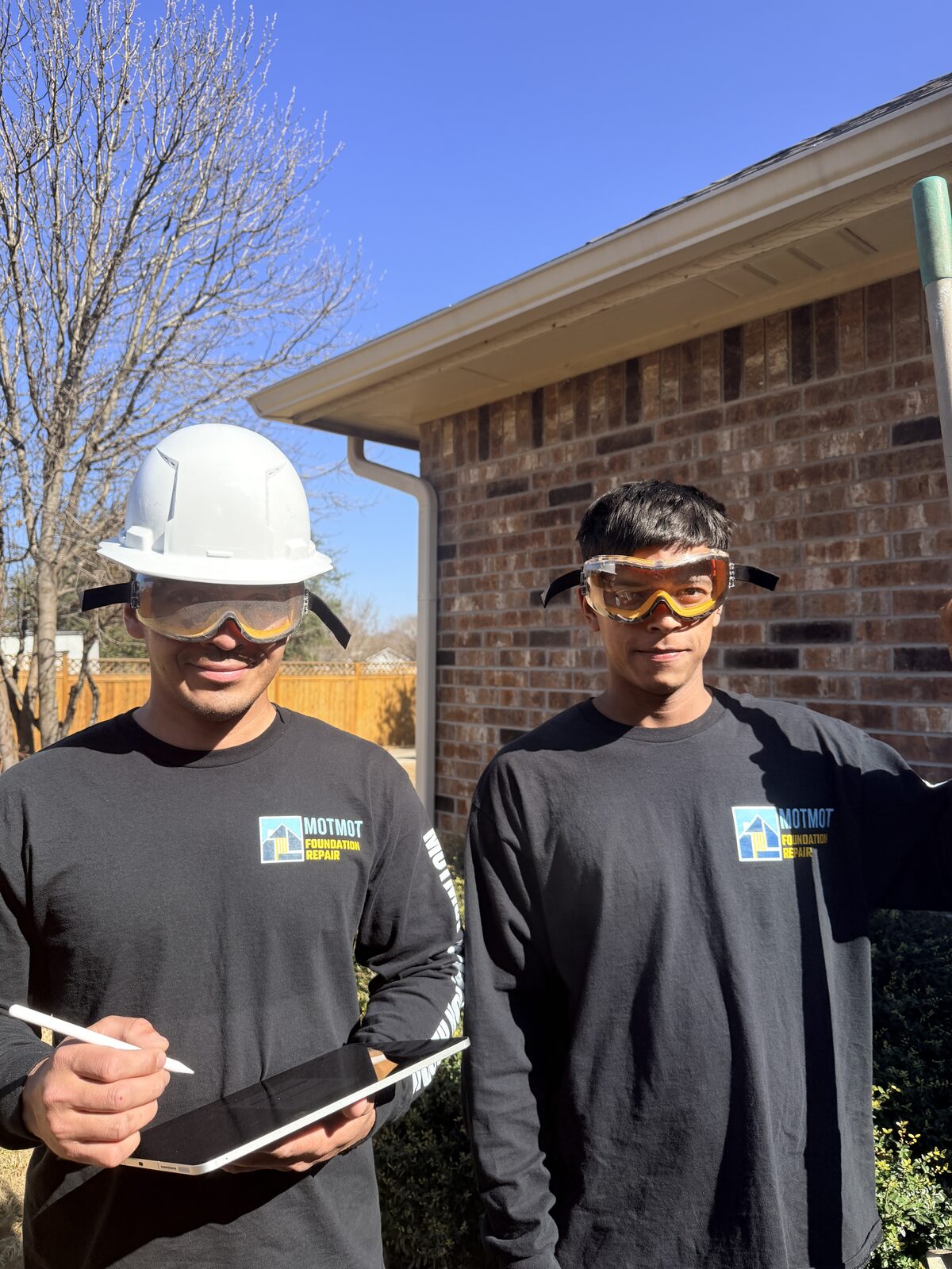 Two Motmot Foundation Repair technicians in safety gear outside a brick home in Central Texas