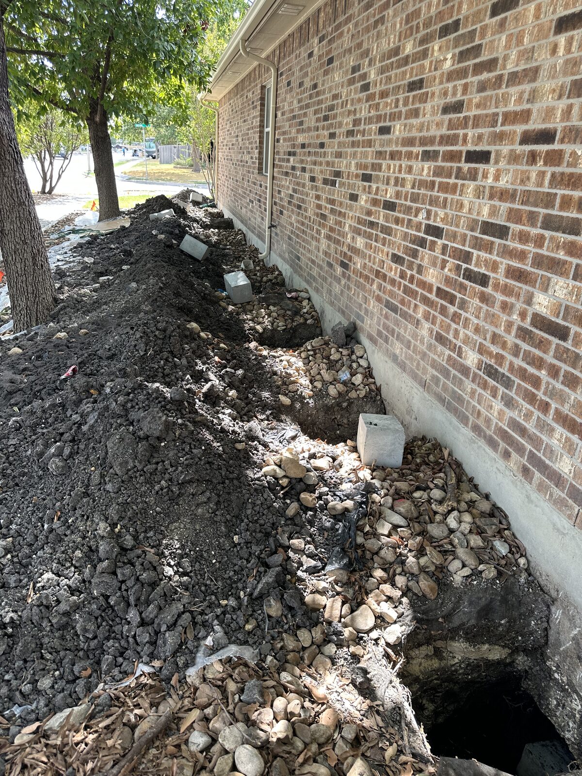 Open trench with pier pits dug through a river-rock bed along a brick home's foundation