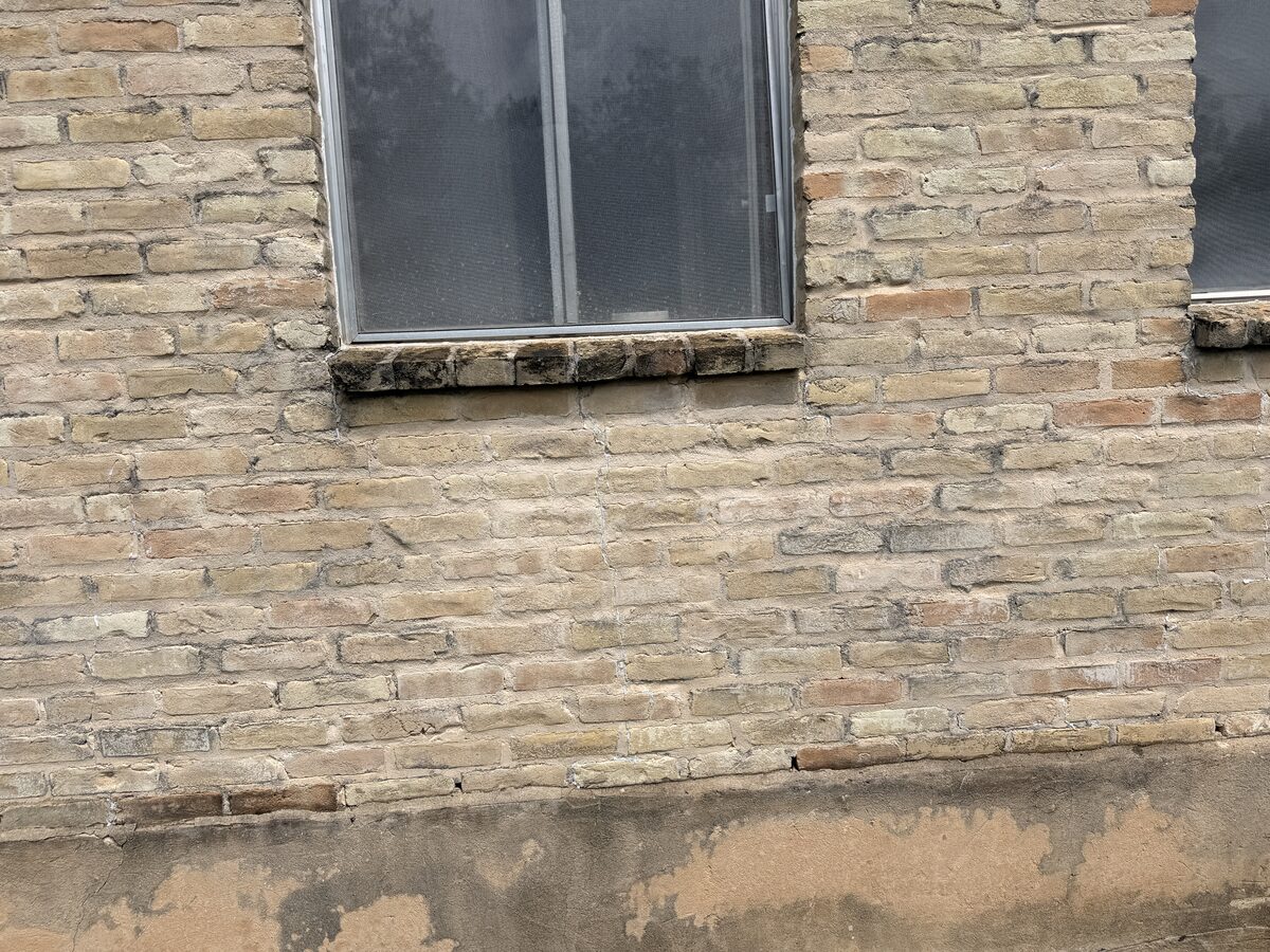 Patched mortar and new cracking at the base of a brick wall below a window, showing ongoing foundation movement