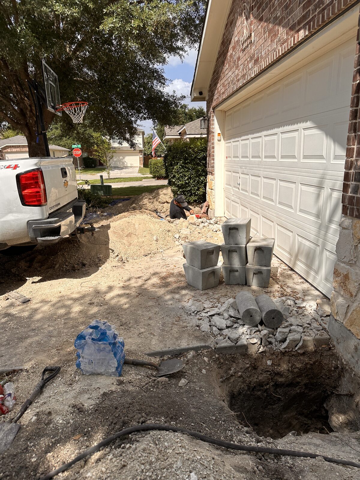 Stacked concrete pier cylinders and an open pit with hydraulic hoses beside a garage during foundation leveling