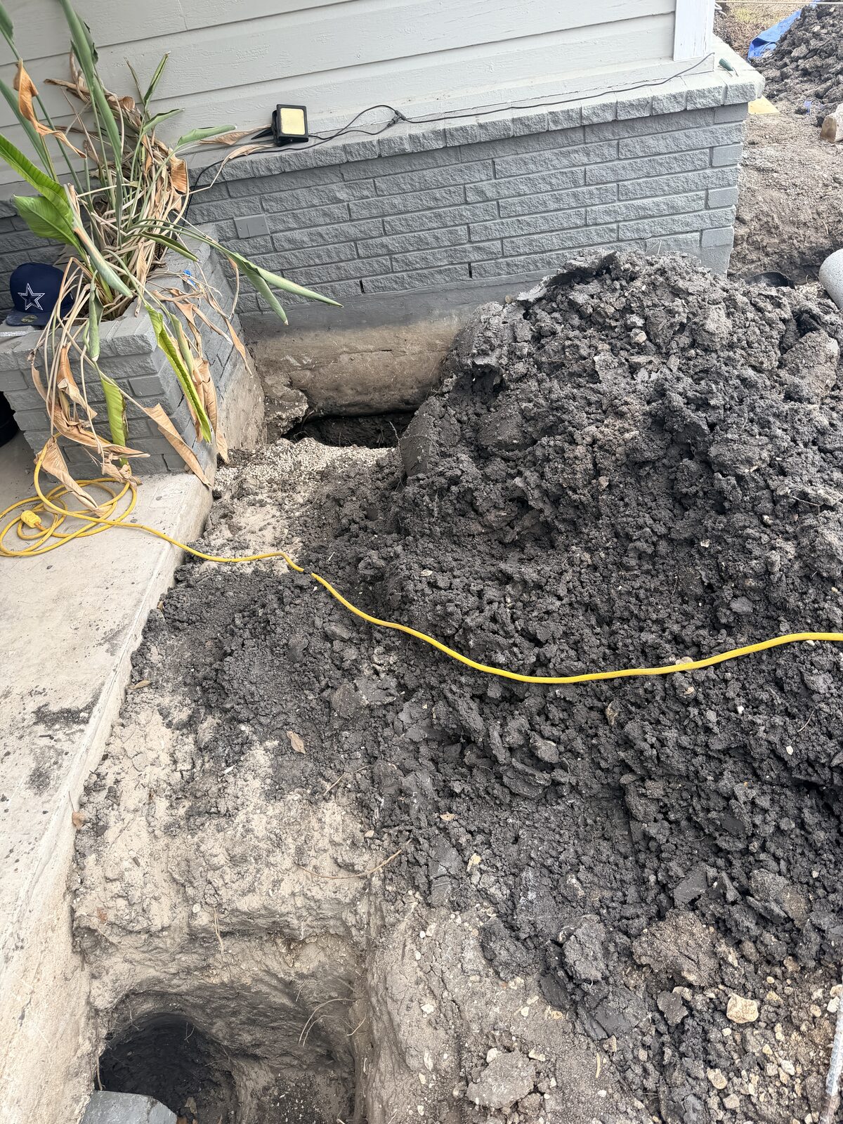 Excavated spoil and a pier hole beside a home's stone-pattern skirting during foundation repair