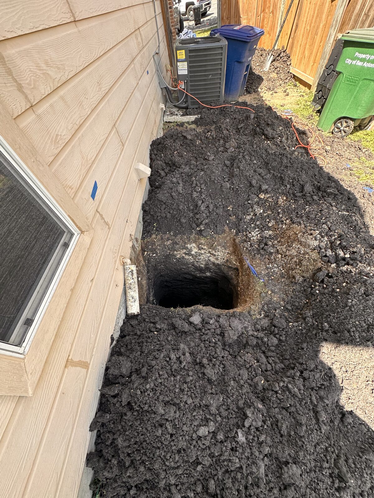 Pier hole dug through dark clay beside a home's slab edge during foundation repair, near the AC unit