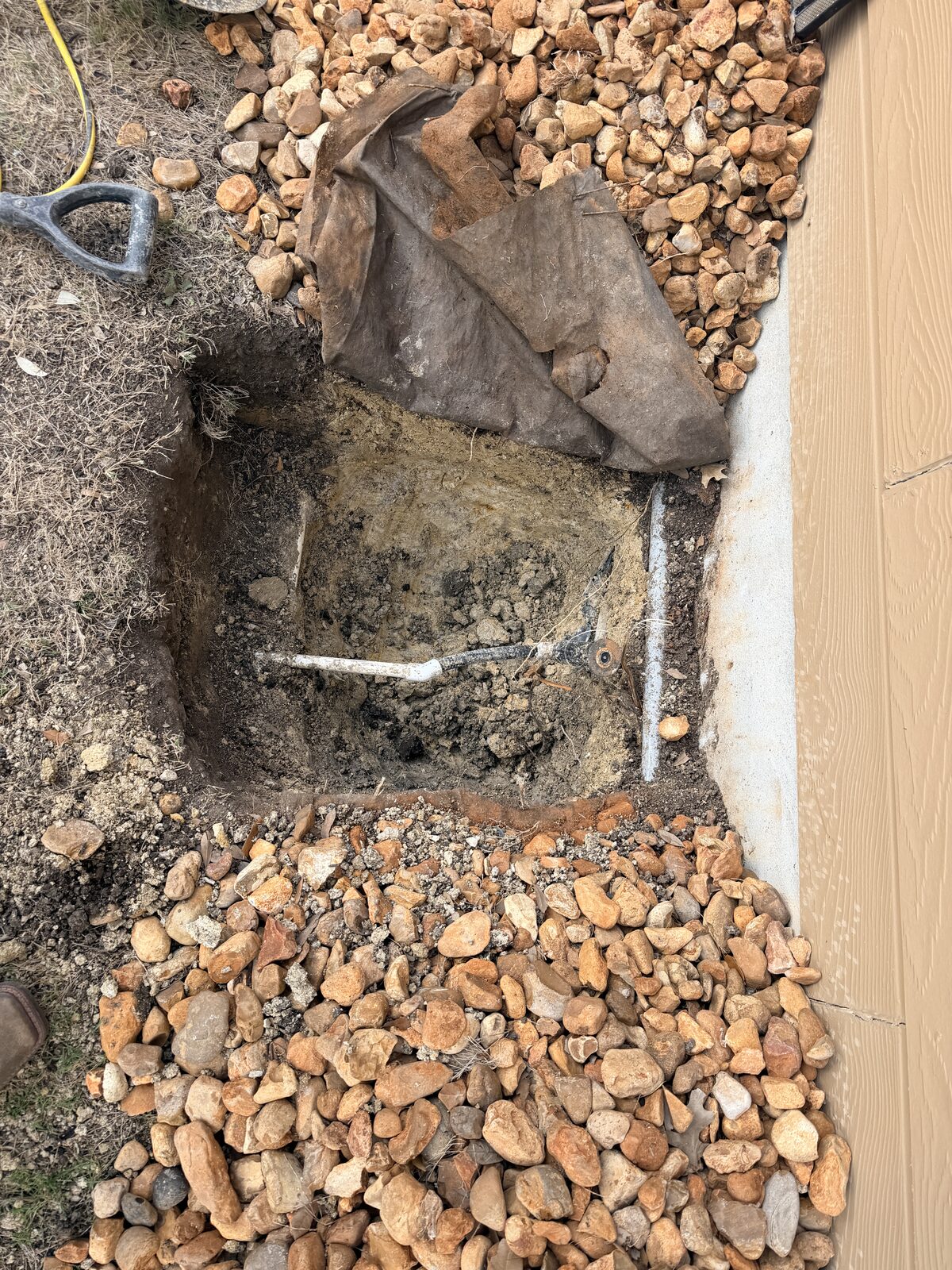 Excavated pier hole at the slab edge with river-rock landscaping pulled back for foundation pier installation