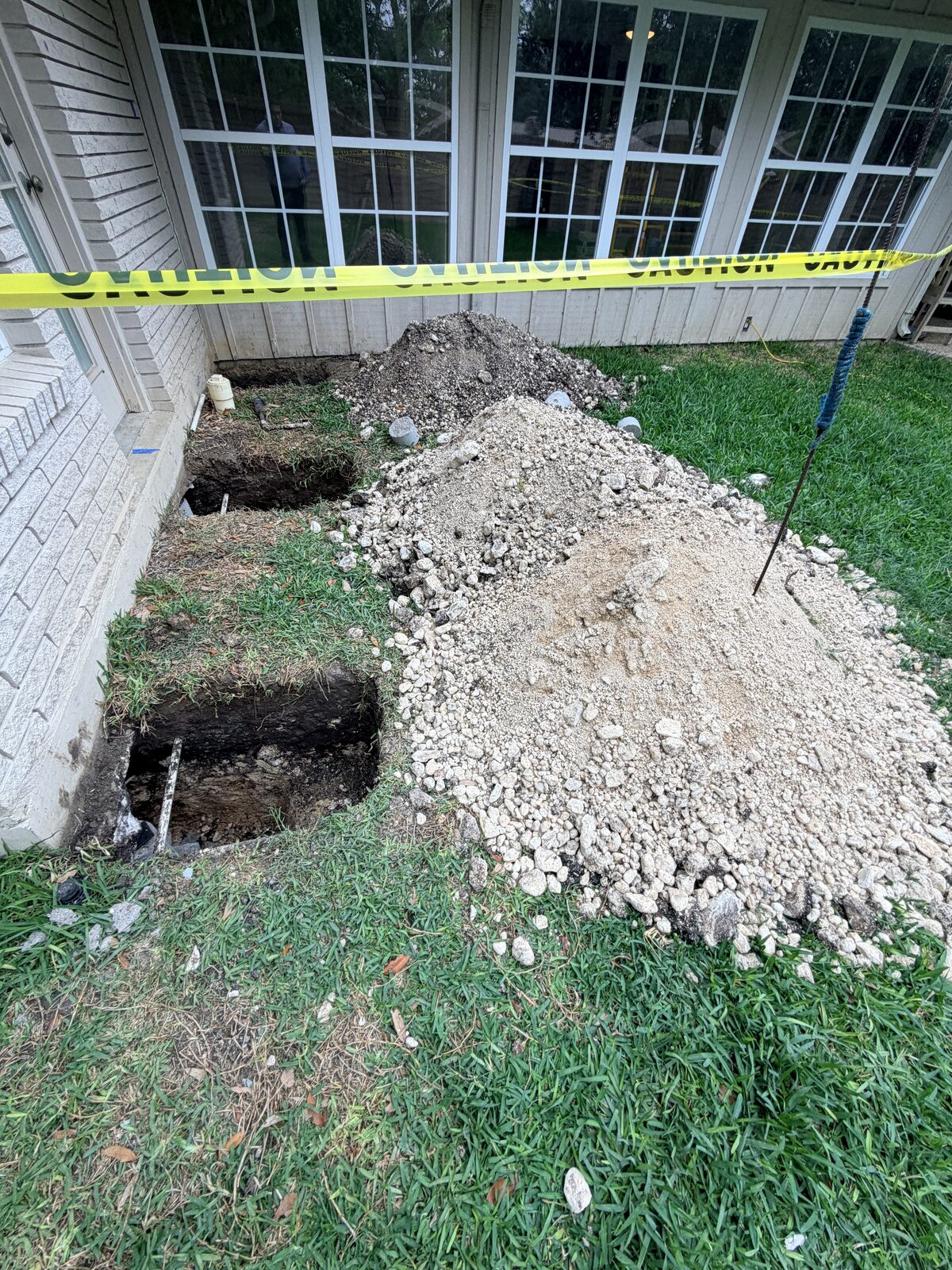 Hand-dug pier holes and excavated soil beside a home's bay window during foundation pier installation