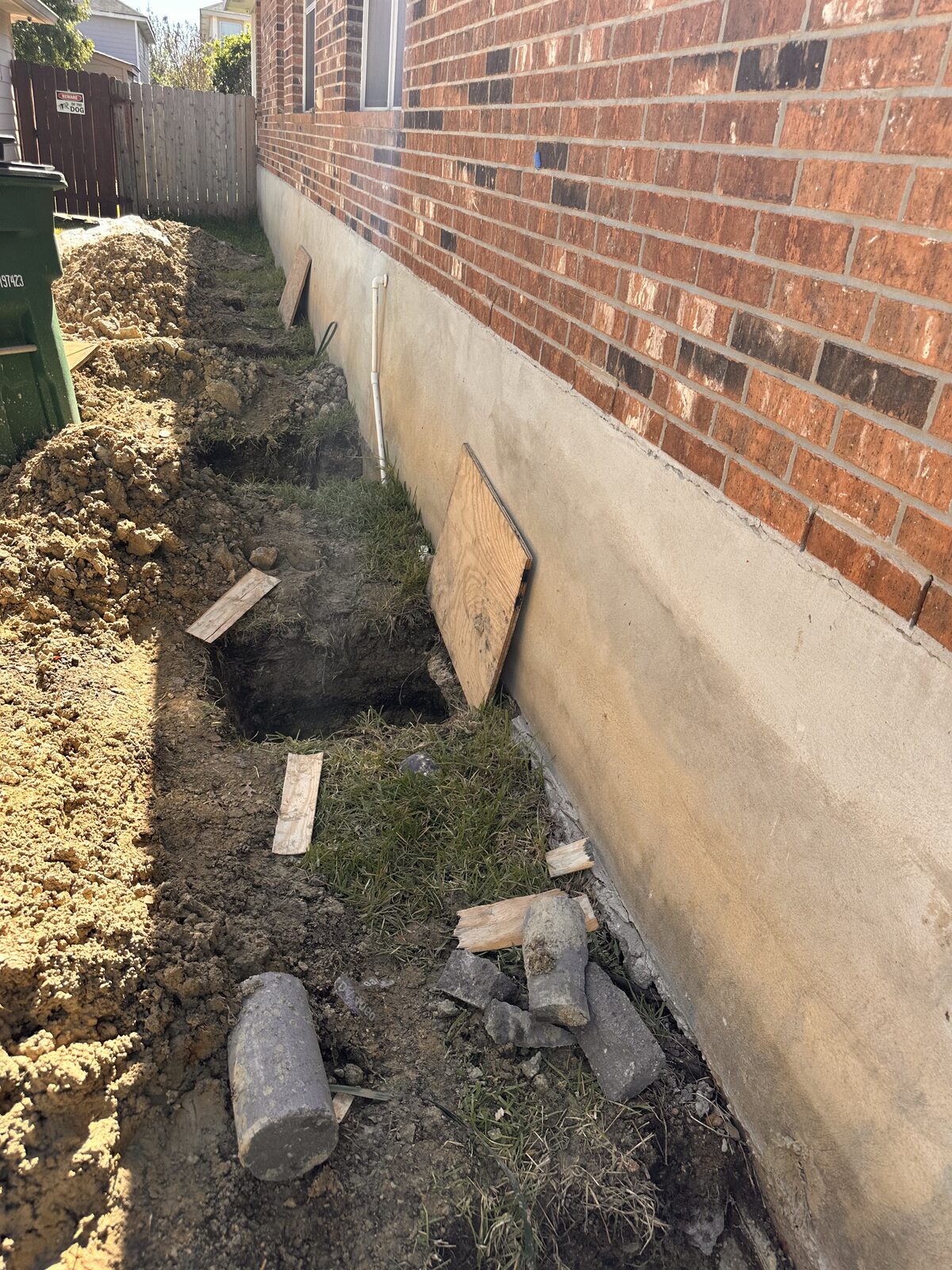 Line of pier holes with walk boards dug along a brick home's stem wall, ready for pilings