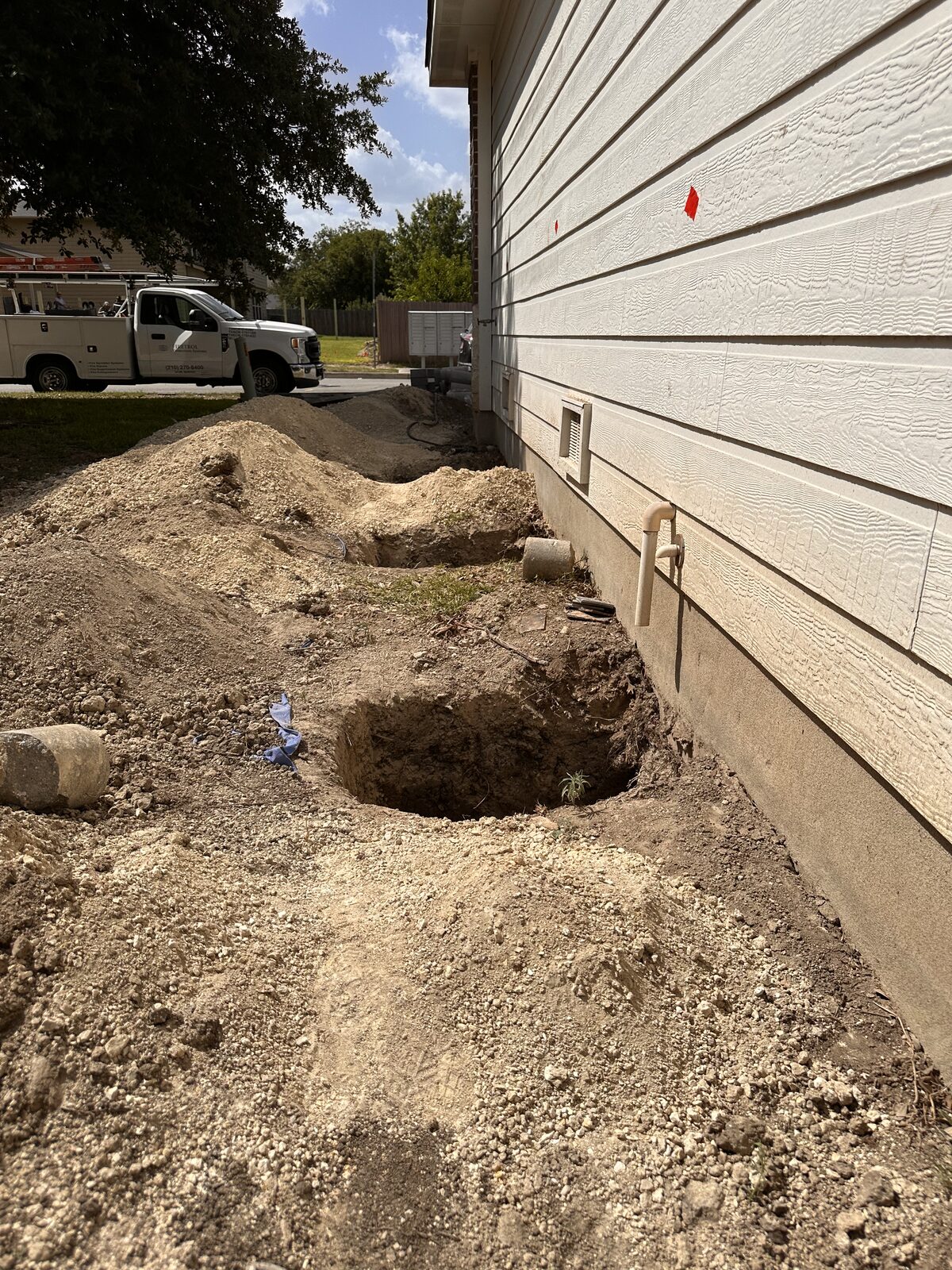 Excavated pier hole and soil spoil along the foundation of a siding-clad home during repair work