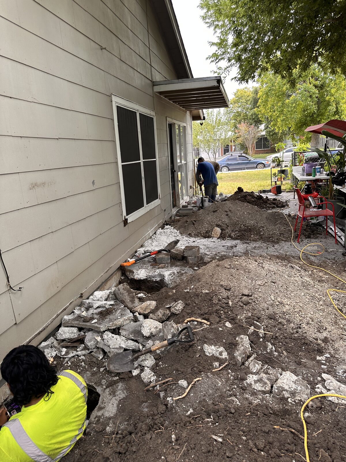 Foundation repair crew working along a cream-sided home, with broken concrete and tools at the job site
