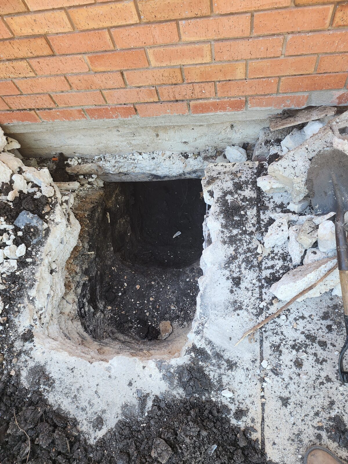 Excavated pier pit beneath the exposed concrete grade beam of a brick home's foundation