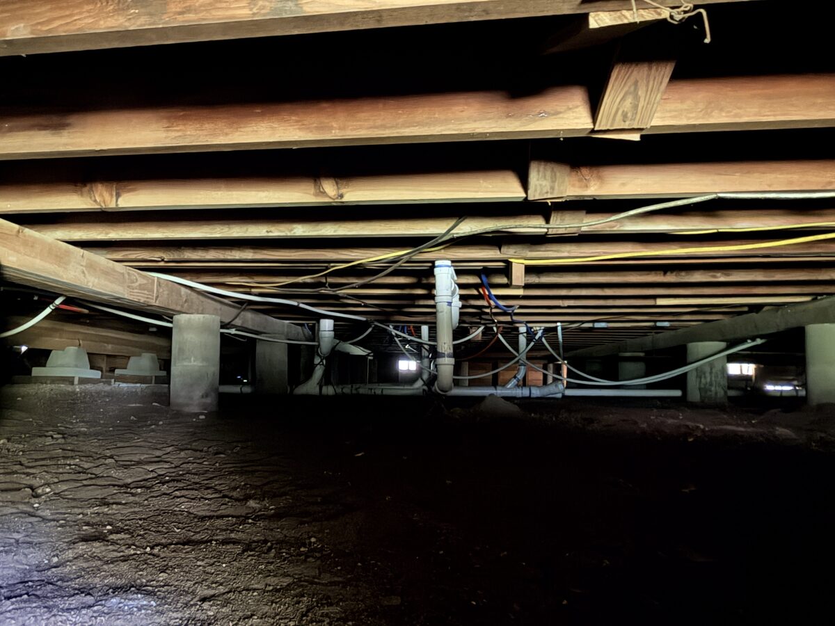 Crawl space view of concrete piers carrying wood beams and floor joists over a dirt floor