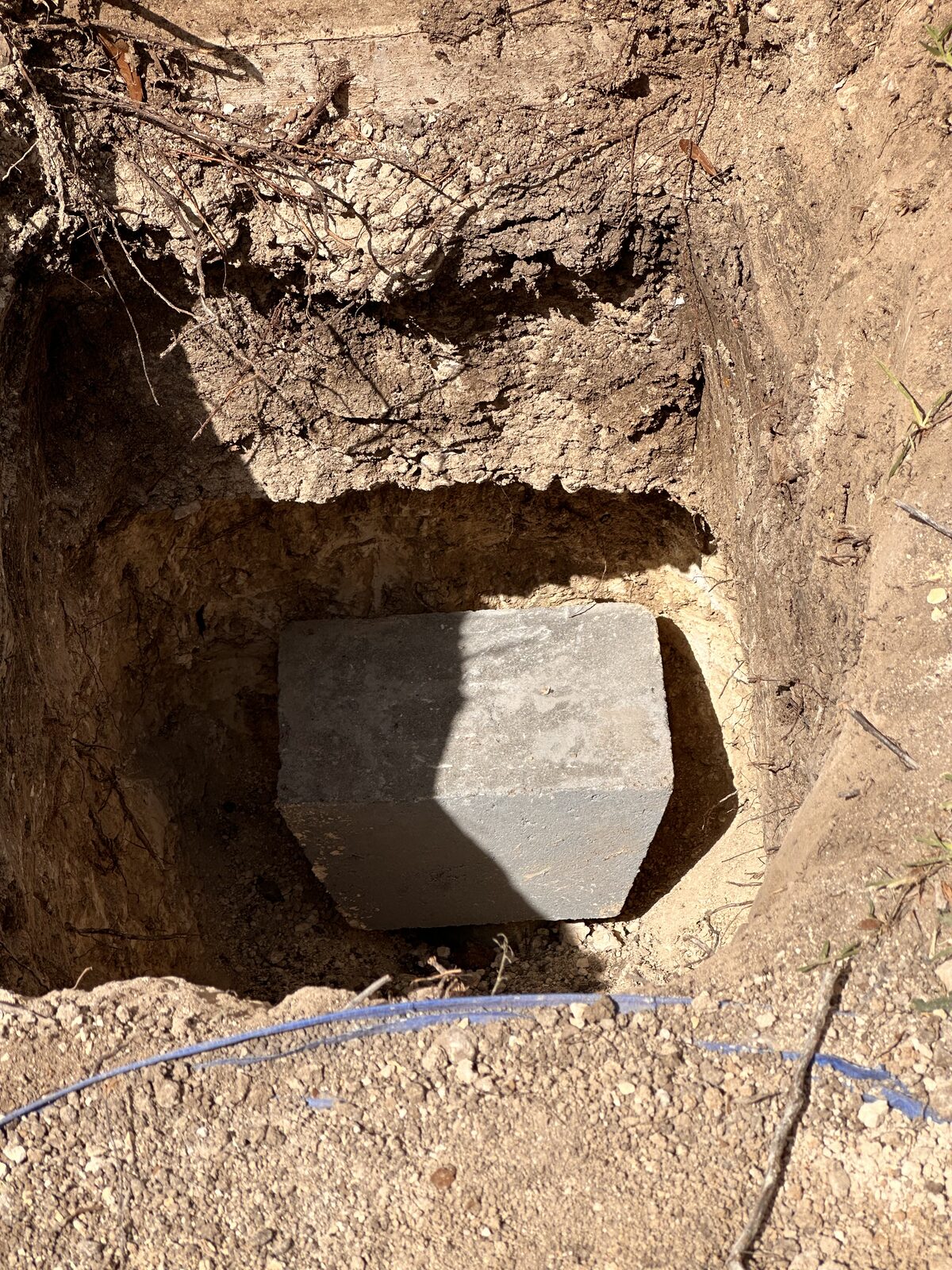 Pressed concrete piling cap in a hand-dug pit under a foundation edge, ready for backfill