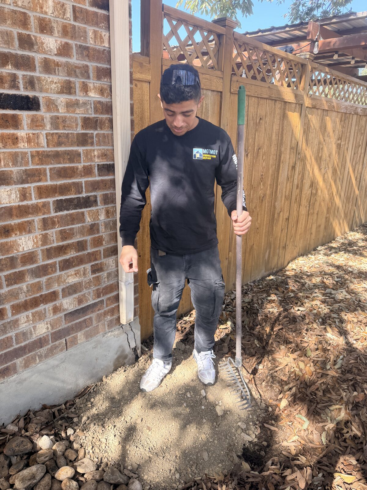 Crew member raking backfill over a completed pier hole beside a brick home's foundation