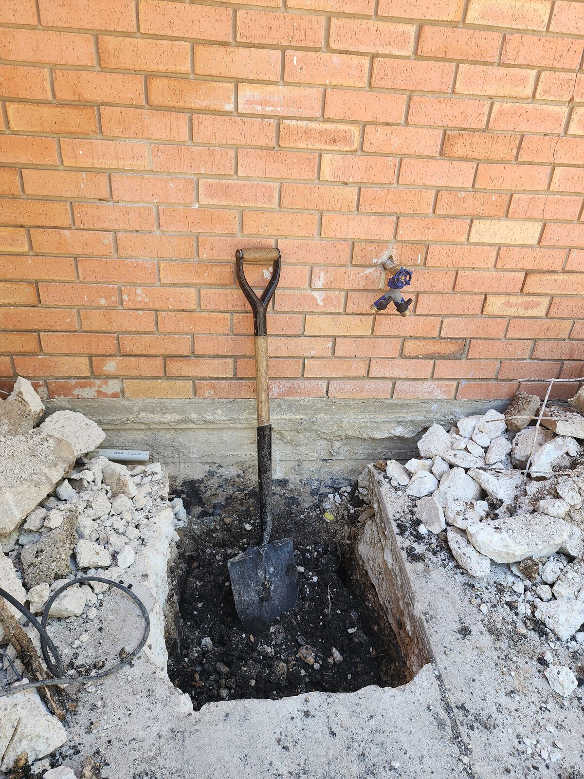 Shovel standing in a freshly dug pier pit beside a brick foundation, with rubble from the exposed grade beam