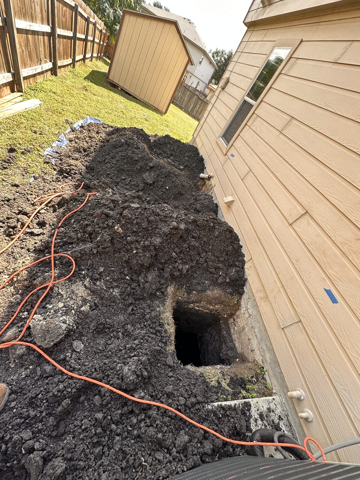 Side-yard excavation with clay spoil piled beside a home's foundation during repair, shed and fence beyond